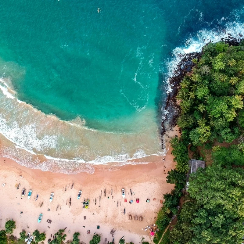 Top-down aerial drone shot of Devinuwara Beach in Matara, Sri Lanka, showing turquoise ocean waves crashing on a sandy shore lined with local fishing boats and palm trees.