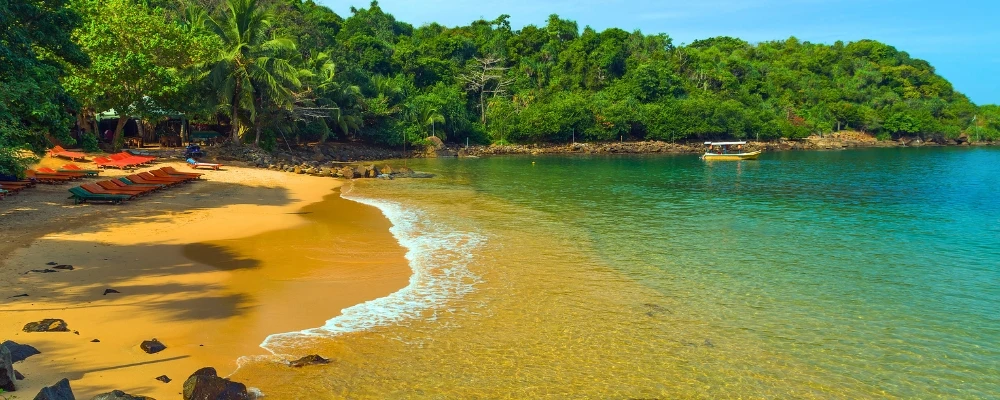 A wide panoramic view of Jungle Beach in Unawatuna, showing golden sand lined with orange sunbeds, clear turquoise ocean water, a small boat, and a dense green forest hillside under a bright sky near Galle, Sri Lanka.