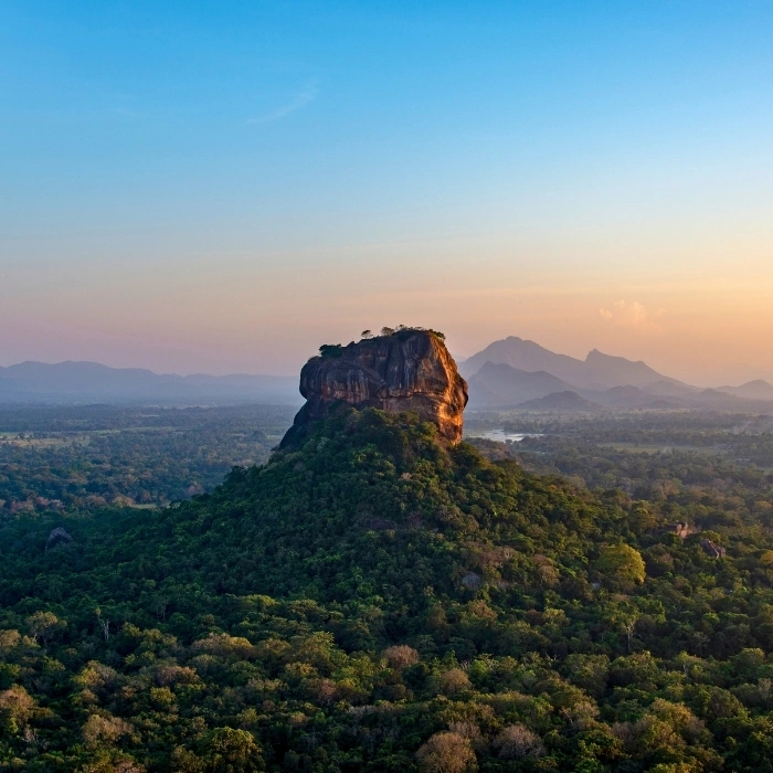 An elevated, high-angle panoramic view of the massive Sigiriya Lion Rock citadel rising dramatically out of a dense green forest landscape under a clear sunset sky in Matale District near Dambulla, Sri Lanka.