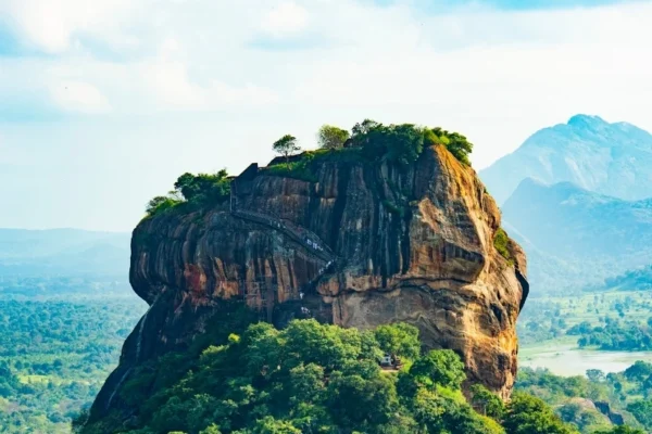 A high-angle aerial view of the Sigiriya Rock Fortress (Lion Rock) in Sri Lanka, showing the ancient monolithic granite citadel surrounded by a dense green jungle canopy under a bright blue sky.