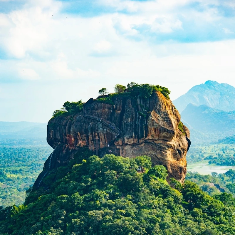 A high-angle aerial view of the Sigiriya Rock Fortress (Lion Rock) in Sri Lanka, showing the ancient monolithic granite citadel surrounded by a dense green jungle canopy under a bright blue sky.