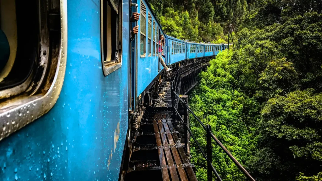 Close-up side view of a passenger hanging out of the open door of Sri Lanka's famous blue train as it curves along the historic Demodara Black Bridge over a dense jungle canopy near Ella.