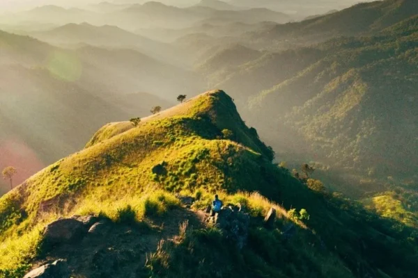 A hiker sitting on the lush green mountain ridge of Little Adam's Peak during a golden morning sunrise, overlooking misty mountain layers in Ella, Sri Lanka.