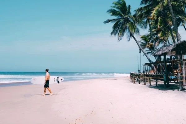 A surfer carrying a surfboard walks along the sunny golden sandy shore at Upali Beach in Arugam Bay, Sri Lanka, surrounded by tropical coconut palm trees and a rustic beach shack—a world-famous surf point for global travelers.